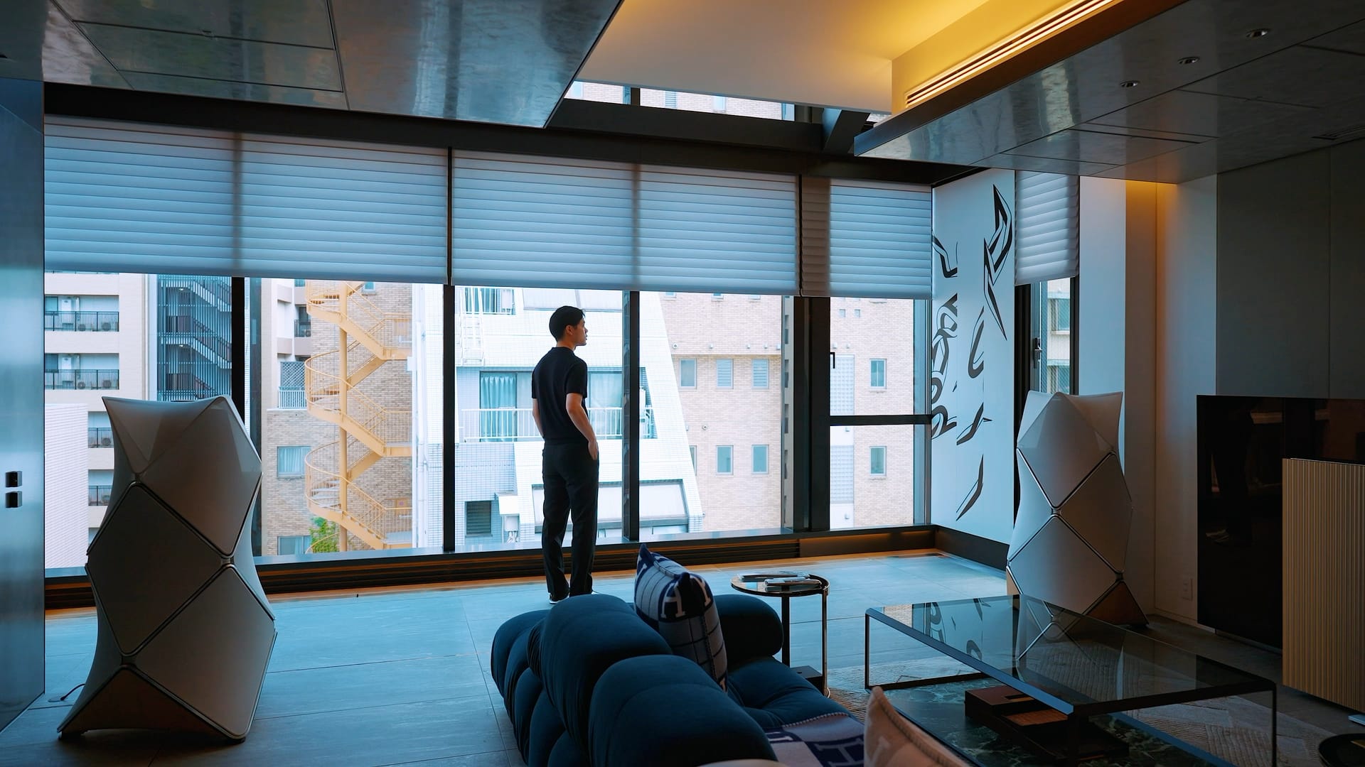 Man looking out floor-to-ceiling windows in a modern living room with automated shades and high-end speakers